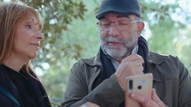 Senior couple standing outdoors in the park, looking at map on smartphone, smiling and discussing the route during walk
