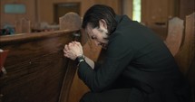 Young, emotional, anxious, and stressed man with long hair and black suit sitting in old church in worship and praying.