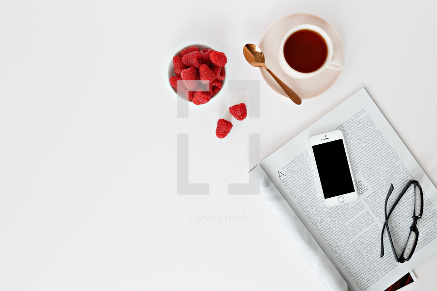 Raspberries in a bowl, tablet, magazine, reading glasses, and coffee cup 