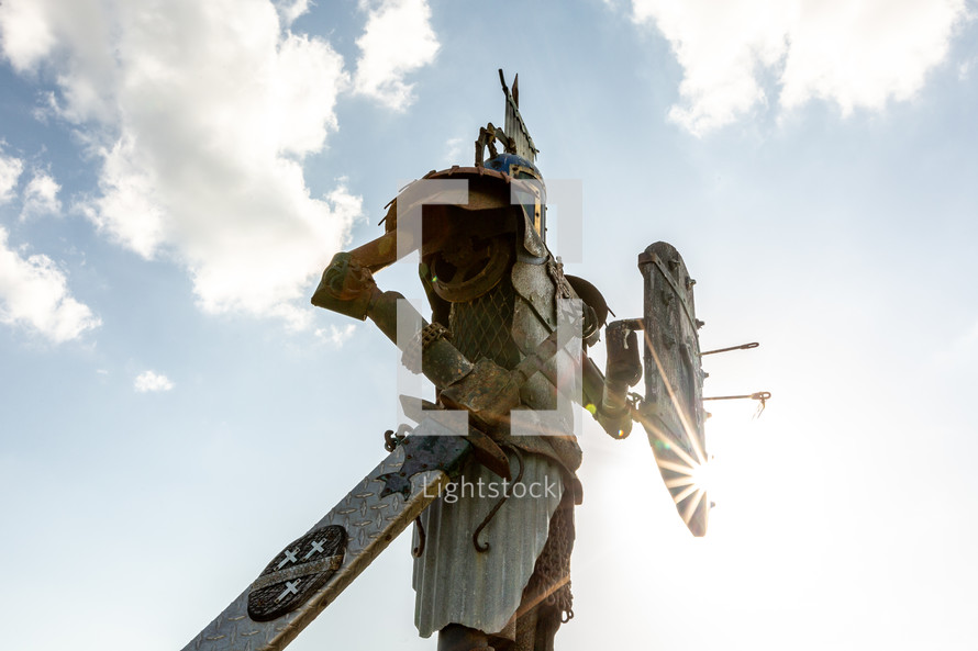 Statue of knight in metal armor holding sword and shield with sky background