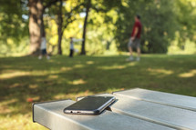 cellphone on a picnic table and a family playing at a park 