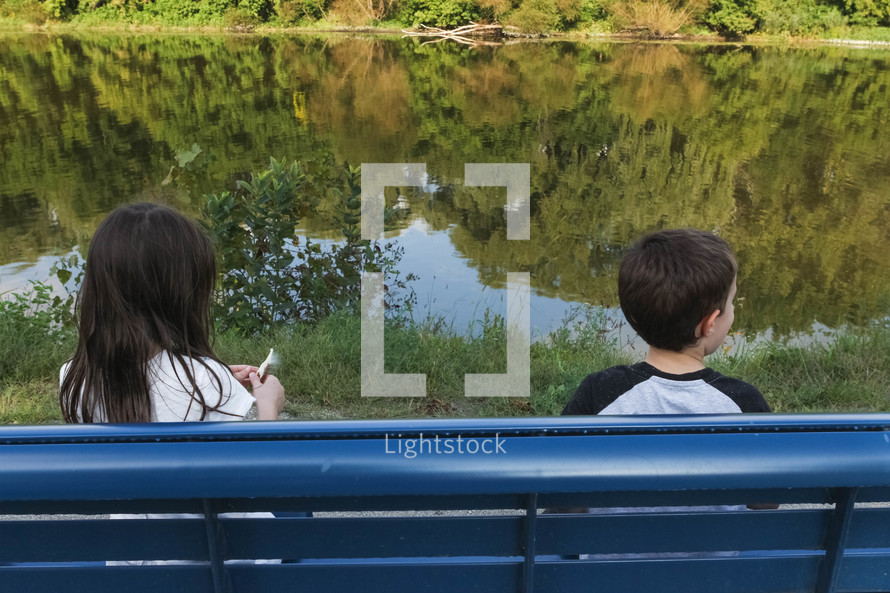 children sitting on a park bench looking out at a pond 