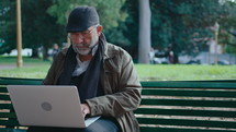 Elderly man with gray beard sitting on bench in the park, typing on laptop, working online outdoors. Medium shot with copy space
