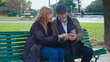 Senior wife and husband sitting together on a bench in the park, checking social media on their smartphone and having a talk, spending the day outdoors. Medium long shot
