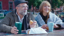 Beautiful senior woman smiling and talking with husband, sitting outdoors at table in the park, drinking coffee from travel mugs and eating pastry
