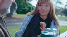 Positive senior couple sitting outdoors in the park, drinking coffee from travel mugs, eating pastry, smiling and chatting. Rack focus shot
