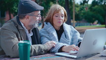 Senior couple sitting outdoors at table in the park, surfing the Internet on laptop and having discussion. Medium shot
