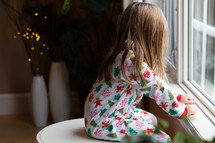 child in Christmas pajamas looking out a window 