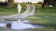 Jesus Christ walking on a country road with a pond reflection.
