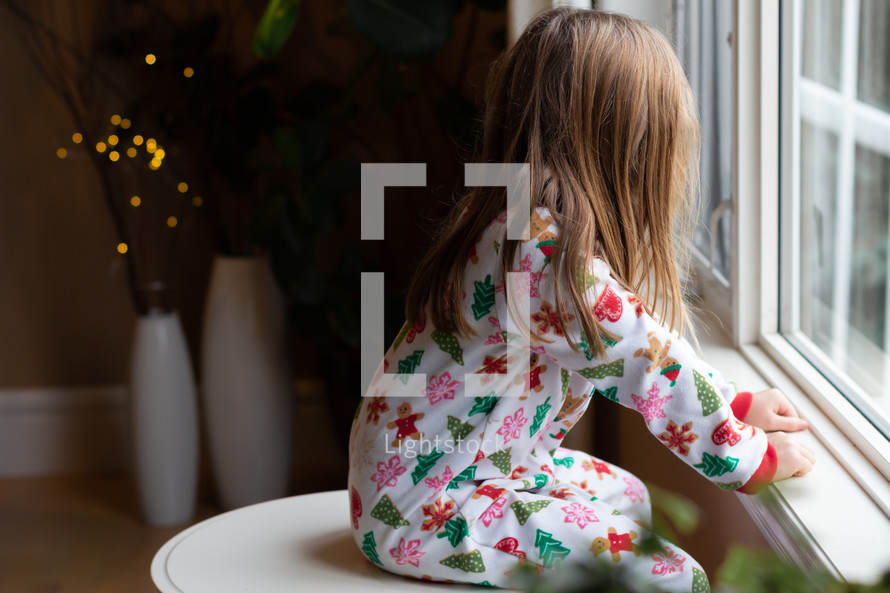 child in Christmas pajamas looking out a window 