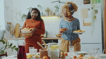 Two young Black women serving dishes on table, smiling and chatting while preparing for family dinner at home
