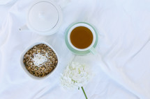 bowl of oatmeal, tea pot, cup of tea, and flowers on sheets on a bed 