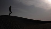 Man walking in middle eastern desert sunset in a desert nature landscape. Sand blowing in wind over desert sand dunes in evening desert sunset, sunlight in cinematic slow motion.