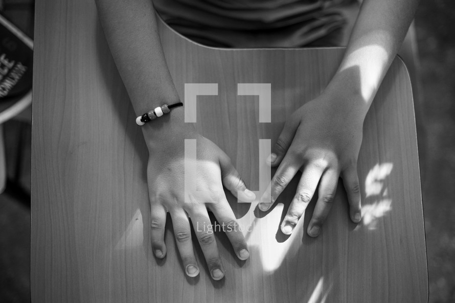 Black and white photograph of a kid's hands on a school desk wearing a gospel bracelet.
