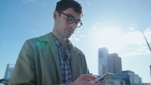 Young handsome businessman with painted nails standing outdoors on sunny day, using smartphone, taking photos of cityscape. Medium shot, low angle view
