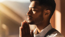 Indian man praying with closed hands with a bright light shining on him close to a window.