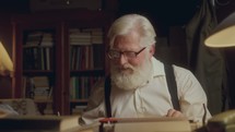 Elderly writer with white hair and beard using typewriter and reviewing papers while working on his book in cozy study with warm light
