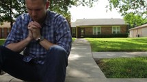 Man sitting on a curb in front of mailbox and house in prayer.