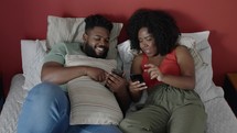 Happy young couple in bed on their phone on social media, browsing internet and online meme. Black couple in bedroom with smartphone. Love, relationship and relax 