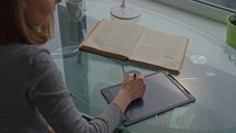 Woman reading book indoor making notes using digital tablet sitting at the desk smart education