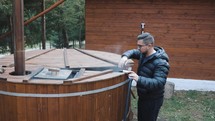 Man Opens Lid Of Big Wooden Hot Tub And Mixes Water With A Mixing Oar. medium shot