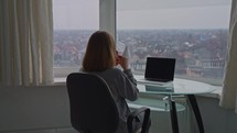 Woman sitting in front of big window enjoying coffee in home office.