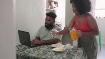 black male freelancer  working online at laptop. Wife support and serve juice to his husband. Online education, work concept.