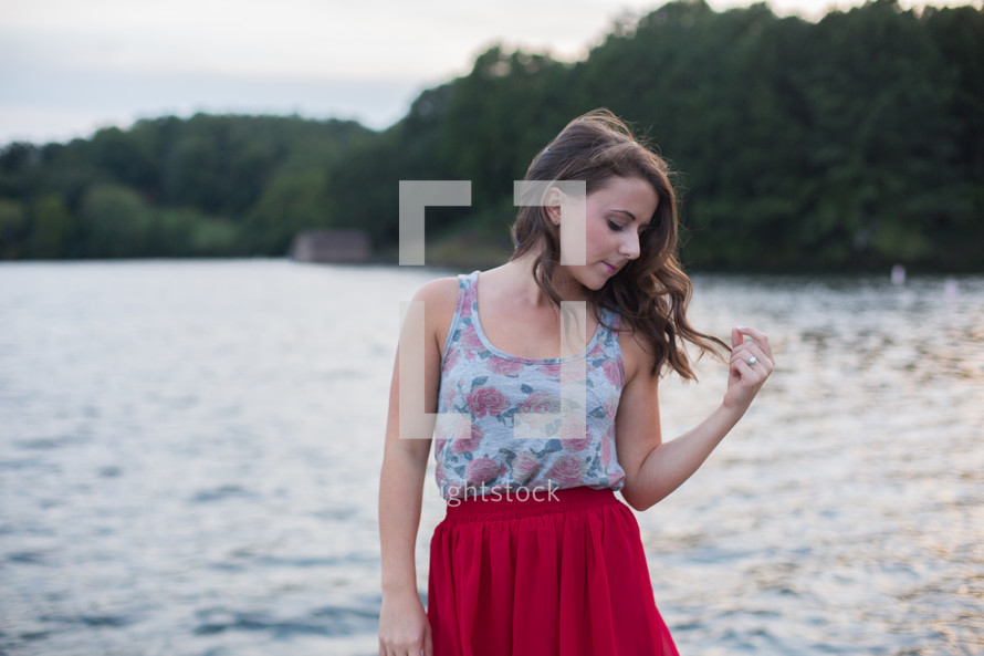 a brunette touching her hair standing in front of a lake 