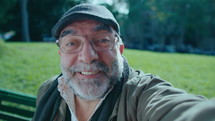 Cheerful senior man with gray beard looking at camera, speaking and showing thumb up, making a video call outdoors in the park. POV shot
