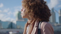 Young curly-haired African-American businesswoman standing outdoors in downtown, looking at cityscape, scrolling on smartphone. Medium close-up shot
