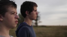 Two happy, smiling young men, best friends, standing in nature watching the summer sunset in cinematic slow motion. 