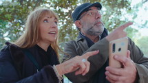 Senior couple standing under the tree in the park, using map on smartphone, discussing the route when having walk outdoors
