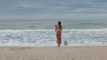 Mother walking on tropical beach as son goes swimming - wide shot from behind