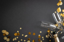 Champagne bottle, champagne flutes and gold streamers and confetti on a black background with copy space
