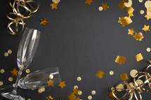 Two champagne flutes on a black background with gold streamers and confetti, with copy space