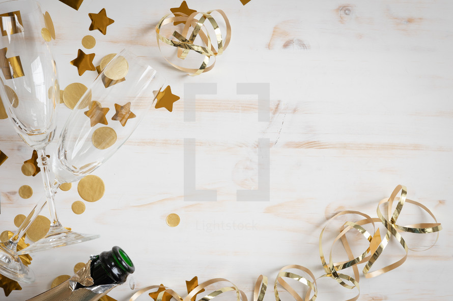 Border of Champagne bottle, two glasses and gold streamers and confetti on a white wood background with copy space