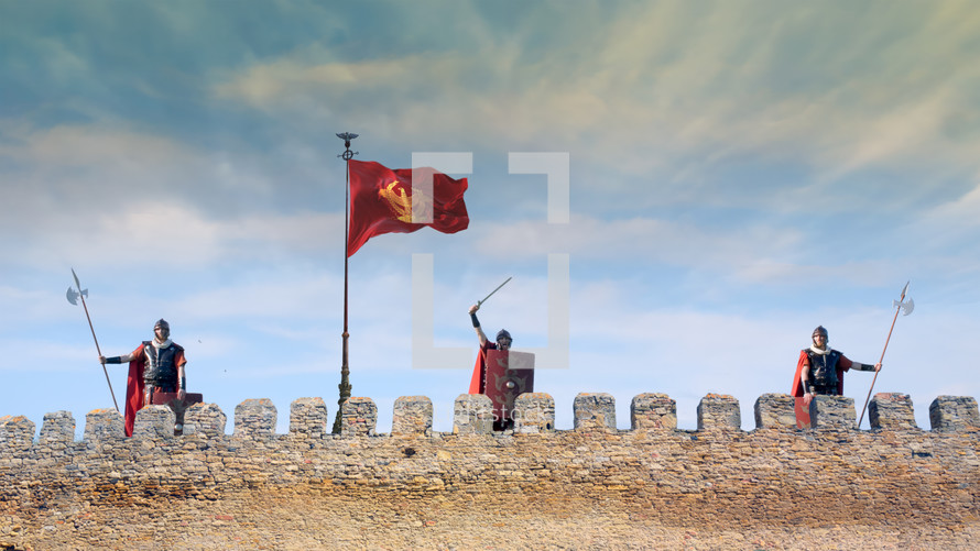 Three soldiers standing on a fortress wall with the flag of the Roman Empire on a cloudy day.
