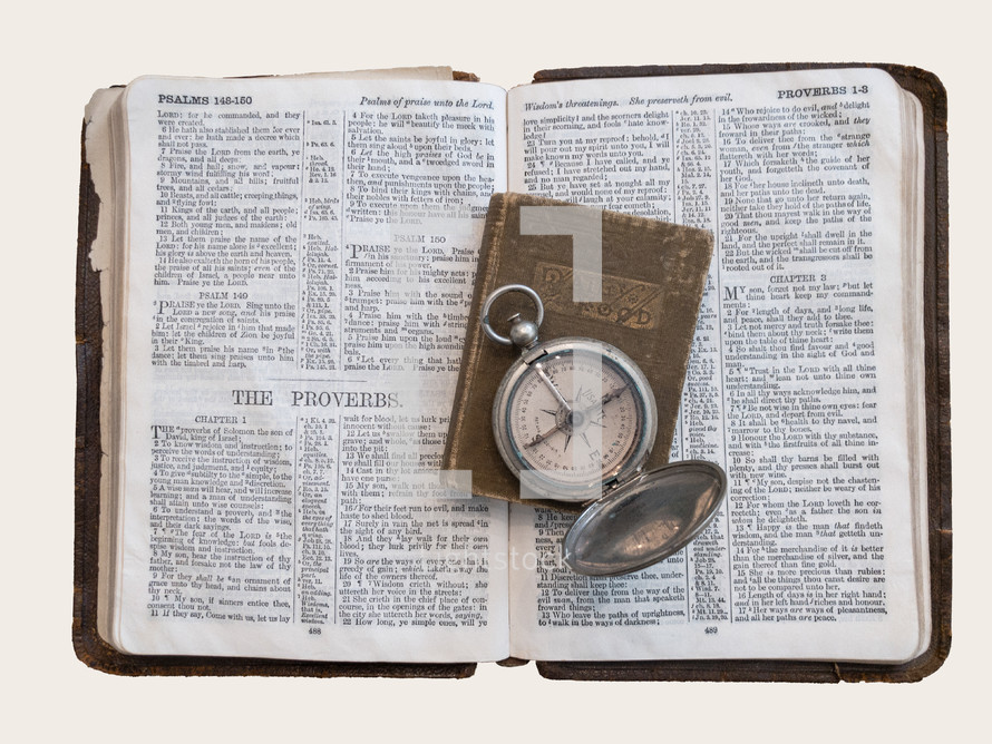 three well-worn, antique items: an open compass rests atop a small devotional book on a larger book open to the end of the Psalms and the beginning of the Proverbs