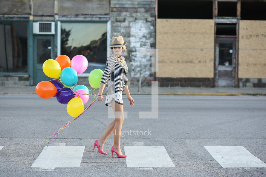 Woman in a hat and high heels walking down the street holding balloons.