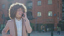 Young African American businesswoman standing outdoors on street in the city, smiling and speaking during phone call. Medium shot, lens flare
