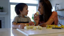 Mother and toddler son eating bananas on kitchen counter - smiling and laughing