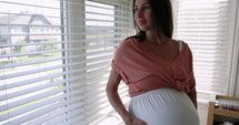 Pregnant woman relaxing looks out her bedroom window in morning