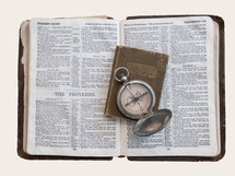 three well-worn, antique items: an open compass rests atop a small devotional book on a larger book open to the end of the Psalms and the beginning of the Proverbs