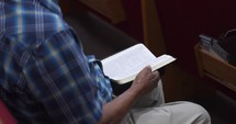 Over the shoulder view of a man sitting in a church pew flipping through a Bible.