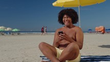Cheerful african american young man hold mobile phone using app, social network, video call, watching funny videos and laughing sitting at the beach lifestyle. Concept online browsing film.