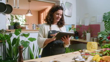 Mid-aged woman in apron standing by kitchen table with food ingredients, using digital tablet and searching for video recipes on the Internet before cooking dinner at home
