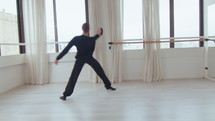 Young male ballet dancer holding one leg on the barre and stretching hamstring muscles while warming up in the studio. Full shot, zoom-out
