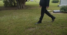 Sombre, sad young man in black suit walking in cinematic slow motion in cemetery through graveyard tombstones.