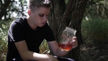Young man, teenage boy with black eye sitting outside in trees drinking alcohol from jar. 