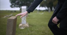 Young, sad man in black suit kneeling in cemetery at graveyard tombstone grieving in cinematic slow motion.